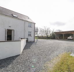 Exterior view of a white two-story house with open doors and gravel driveway with covered parking and cars at Bodegri Cottage Llanrhyddlad near Church Bay