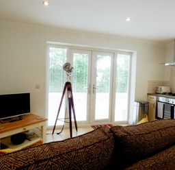 A living room and kitchen area with a sofa, tripod floor lamp, TV on a wooden table, and kitchen appliances at The Hideaway in Verwood