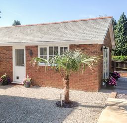 A small brick building with a white door and windows surrounded by gravel and a palm tree at The Hideaway in Verwood