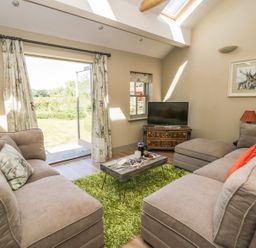 A living room with two grey sofas a green rug a wooden coffee table a television on a cabinet an open door with curtains and a window at Providence Cottage in Easingwold