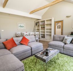 A living room with gray sofas and orange and patterned cushions a wooden coffee table on a green rug and a dining table with white chairs and bookshelves at Providence Cottage in Easingwold