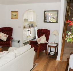 A living room with two red armchairs a white sofa a wooden side table a lamp flowers on a table and stairs with a kitchen visible through an archway at Coachmans Cottage in Witherslack