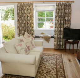 A living room with a beige sofa and floral cushions a patterned rug curtains on windows a television on a wooden table and a basket with logs at Coachmans Cottage in Witherslack