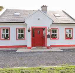 A white house with a red door and window frames surrounded by greenery at Lough Mask Road Fishing Cottage in Ballinrobe County Mayo