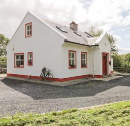 A white house with red trim around windows and door a bicycle leaning against the wall and trees in the background at Lough Mask Road Fishing Cottage in Ballinrobe County Mayo