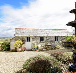 A stone cottage with outdoor chairs and a table in a gravel courtyard with a tiered fountain and plants at The Snug Rescorla near St Austell