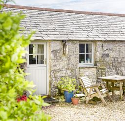 A stone cottage exterior with a white door wooden chairs and a table on a gravel patio at The Snug in Rescorla near St Austell