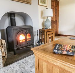 A wood burning stove with fire next to a basket of logs and a wooden table with a book and coasters at Hill Bank in Hutton Roof