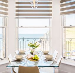 A dining area with a glass table set for four with a loaf of bread grapes and flowers by windows overlooking the sea at No 6 Arlington Villas in Ilfracombe