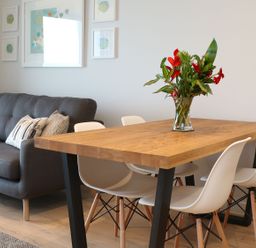 A dining area with a wooden table and white chairs next to a grey sofa with cushions at Little Fistral in Fistral