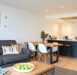 A living area with a gray sofa and patterned pillows a wooden dining table with white chairs and a kitchen island with stools at Little Fistral in Fistral