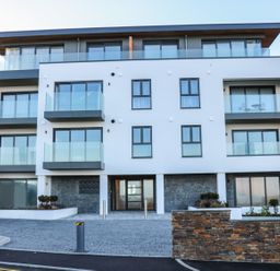 The front view of a modern three-story building with glass balconies and a stone wall at Fistral House in Fistral