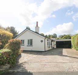 A single story house with a driveway and black garage doors surrounded by hedges and bushes at Glan Dwr in Red Wharf Bay