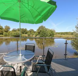 A glass table with green umbrella and black chairs on a deck overlooking a lake with trees at Lakeside Lodge in East Harling near Thetford