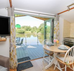 A kitchen dining area with a round table set for two and an open door leading to a deck overlooking a lake at Lakeside Lodge East Harling near Thetford