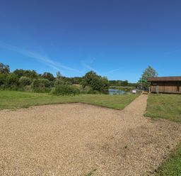 A gravel path leading to a wooden building near a pond with trees and grass at Lakeside Lodge in East Harling near Thetford