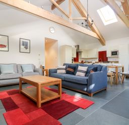 An open living area with two sofas a wooden coffee table a red rug exposed wooden ceiling beams skylight and a kitchen with dining table and chairs at Wheal Grey in Praa Sands