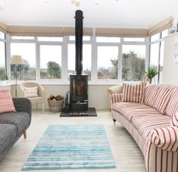 A sunroom with a black stove between a gray sofa and a striped sofa with a blue rug on the floor at The Beach House Criccieth