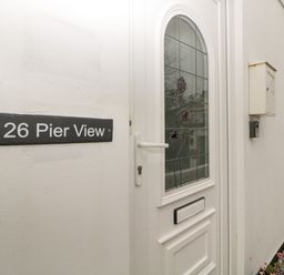 A white front door with stained glass and a house number sign on the wall at 26 Pier View in Paighton