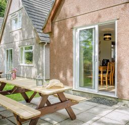 An outdoor patio area with a wooden picnic table and chairs next to a building with sliding glass doors at Dartmoor View in Gunnislake