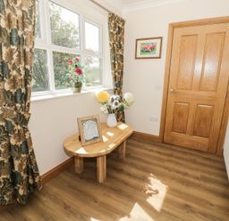 A small room with a wooden table holding flowers and a framed picture near a window with patterned curtains and a wooden door at Rede River Cottage in West Woodburn