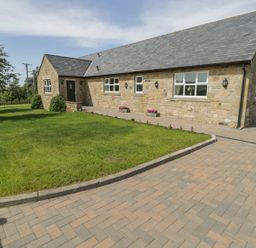 A stone house with a slate roof and green door next to a paved driveway and lawn at Rede River Cottage in West Woodburn