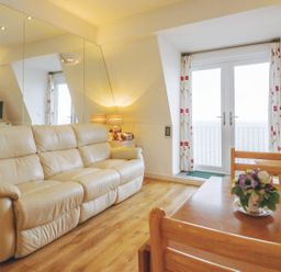 A living room with a beige leather sofa a wooden table with flowers and glass doors with floral curtains at 10 Oceanside in Ilfracombe