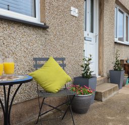 A small patio with a table holding two glasses of orange juice a chair with a green cushion plants in pots and a white door at Gwynfa in Rhyd Uchaf near Bala Gwynedd