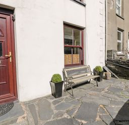 A stone patio with a wooden bench and two potted plants outside a house with a red door at Miners Cottage in Blaenau Ffestiniog
