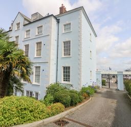 A blue multi-story building with white window frames beside a driveway with greenery and palm trees at Dawn Catcher in Falmouth