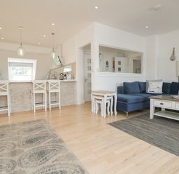 An open living room and kitchen area with a blue sofa, white stools at a breakfast bar, and wooden floor at Dawn Catcher in Falmouth