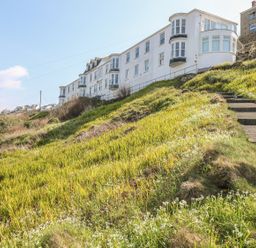 A row of white buildings on a hillside with concrete steps and green grass with small white flowers at Sennen Heights in Sennen Cove near Penzance