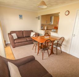 A dining area with a wooden table and four chairs next to a living room with two brown sofas and a kitchen in the background at Surf Hunstanton