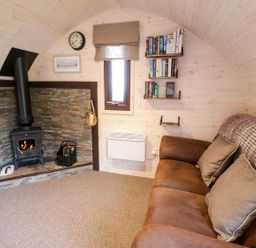 A cozy room with a brown sofa, wood-burning stove, small window, bookshelves, and carpet at Gelert Pod in St Asaph