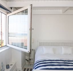A bedroom with an open window showing a view outside and a bed with pillows and a striped blanket at Ner Sea in Marazion