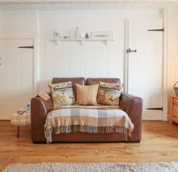 A brown leather sofa with three cushions and a plaid throw in a room with white paneled walls and wooden floor at Ner Sea in Marazion