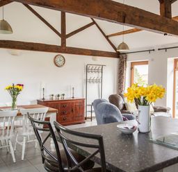 A dining area with white chairs around a wooden table and flowers in a vase a living area with armchairs and large windows at 1 Beacon View Barn in Beguildy near Knighton