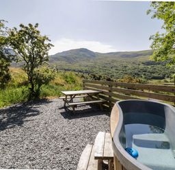 An outdoor area with a hot tub and a wooden picnic table with benches surrounded by a wooden fence and trees at Moelfre - Shepherds Hut in Llanbedr