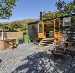 A black wooden shepherds hut with open doors leading to a kitchen area outside seating and a wooden hot tub at Moelfre - Shepherds Hut in Llanbedr