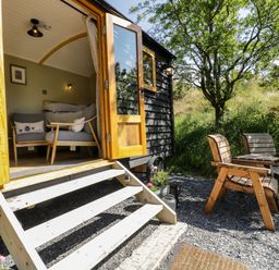 A small cabin with open doors showing two chairs inside and two wooden chairs outside near trees at Moelfre - Shepherds Hut in Llanbedr