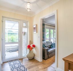 An entryway with a glass door leading outside a large vase with flowers on the floor and a view into a living room with a leather sofa at Yr Hen Ffald in Pontarddulais