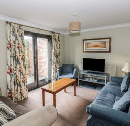 A living room with two sofas a blue armchair a wooden coffee table a flat screen TV on a stand floral curtains and a framed picture at The Lodge off High Street in Hadleigh