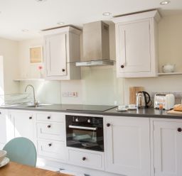 A kitchen with white cabinets, a black countertop, a built-in oven, a stove, a sink with a drying rack, a kettle, a toaster, an egg cup with an egg, a loaf of bread on a cutting board, two cups on saucers, a teapot, a cutting board, a plant, and a fire extinguisher near a wooden door at Salters in Black Dog near Crediton