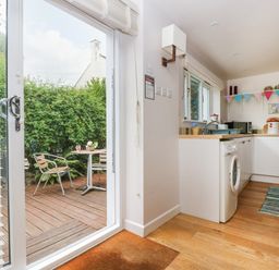 A kitchen with white cabinets and a washing machine near a sliding glass door opening to a wooden deck with two metal chairs and a round table at Bolitho's Retreat in Helford
