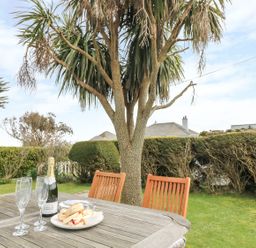 A wooden outdoor table with a bottle of champagne three champagne flutes and a plate with scones in a garden with trees and hedges at Bolenowe in Trebetherick near Polzeath