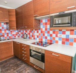 A kitchen with wooden cabinets a tiled backsplash in red blue and light blue colors a built in oven stovetop microwave electric kettle and a double sink at Apartment 2 in Tenby