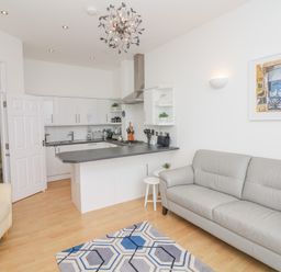 A living room with a gray sofa a cream armchair a patterned rug and a kitchen with white cabinets and a black countertop at Oceans Side in Ilfracombe