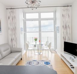 A living room with a gray sofa a round dining table with chairs and a television on a stand at Oceans Side in Ilfracombe