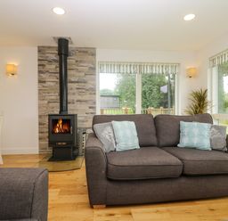 A living room with a wood-burning stove beside a gray sofa with cushions and windows overlooking a garden at Ash in Truro