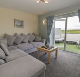 A living room with a gray sectional sofa a wooden coffee table and sliding glass doors leading to an outdoor seating area at Sea Spray in Dinas Dinlle